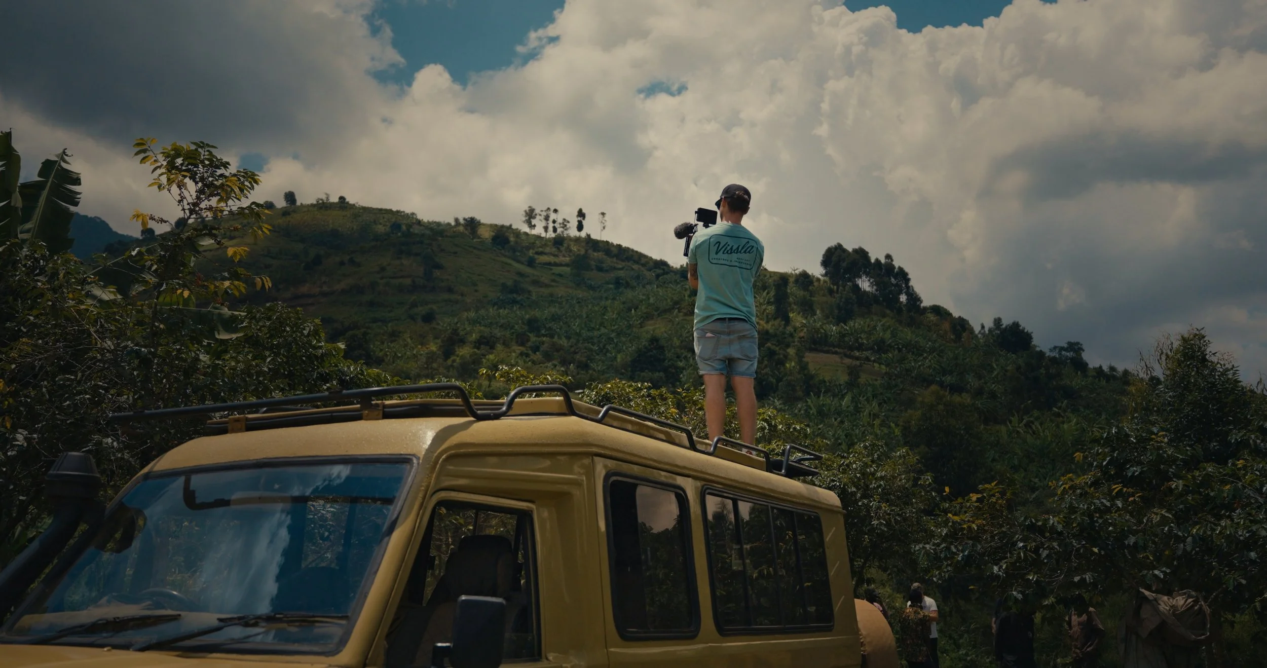 Robin Danehav standing on a safari truck, filming over green hills in Uganda.