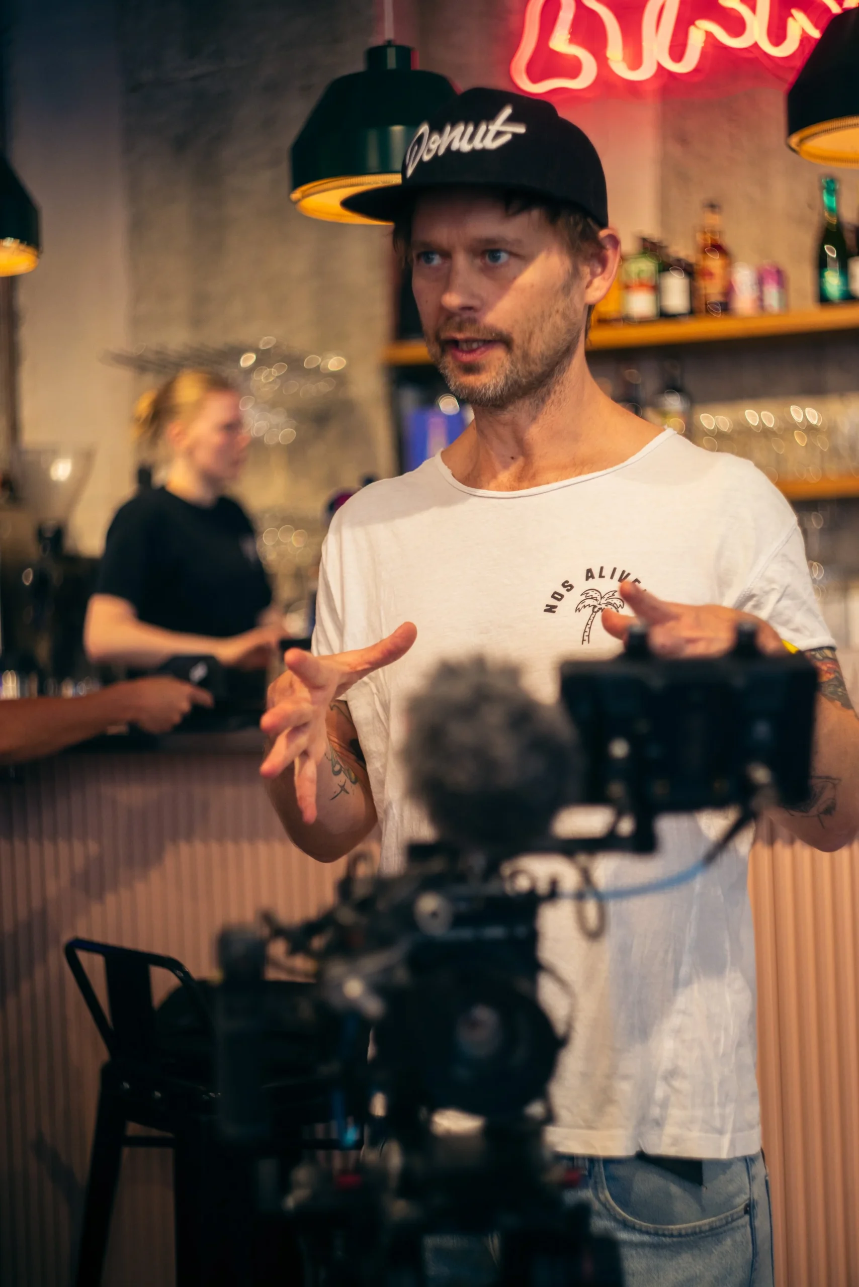 Robin Danehav talking behind a camera in a café, with neon lights in the background.