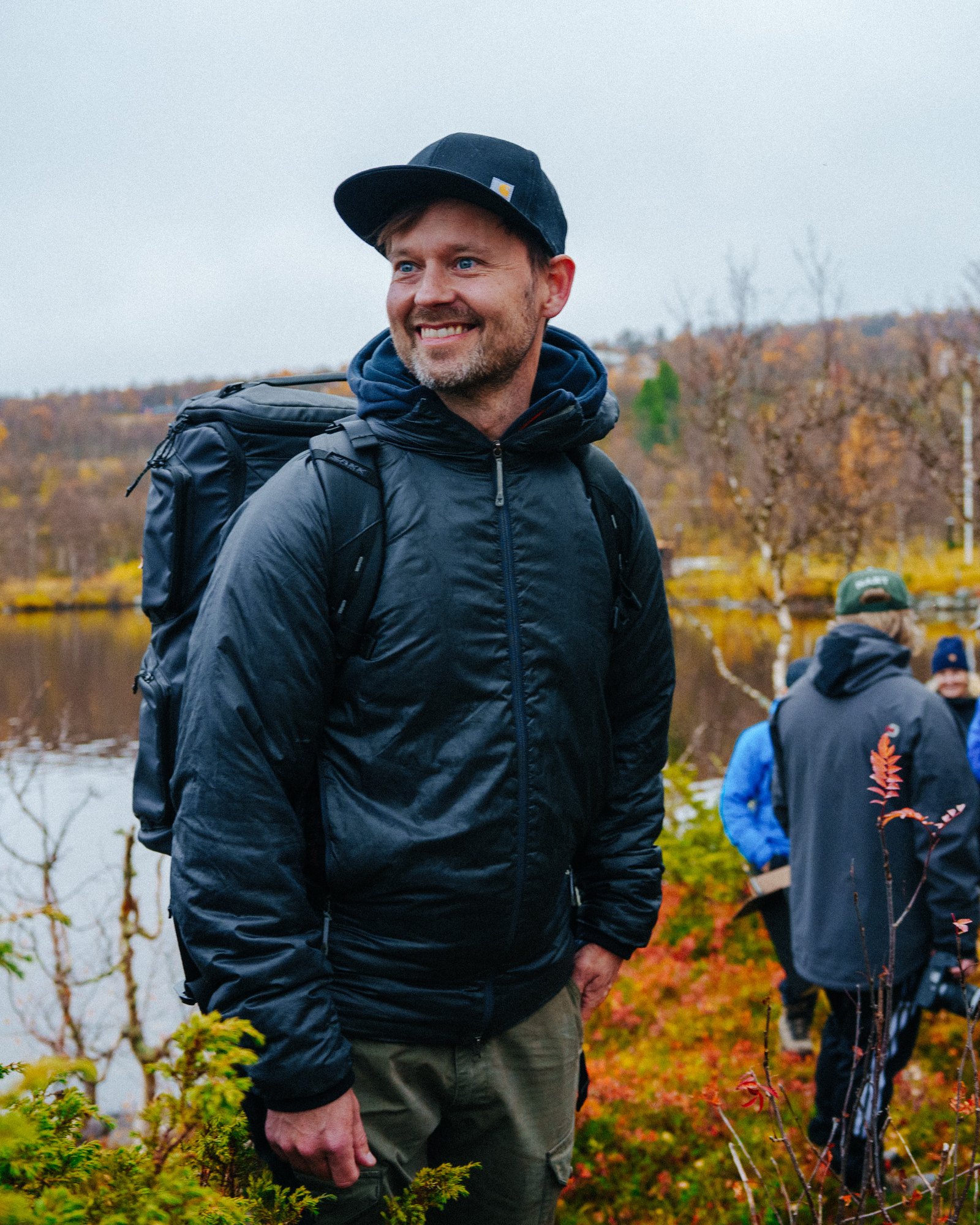 Robin Danehav smiling, in a cap and with a camera backpack, on a shoot by a lake in the Swedish mountains in autumn.
