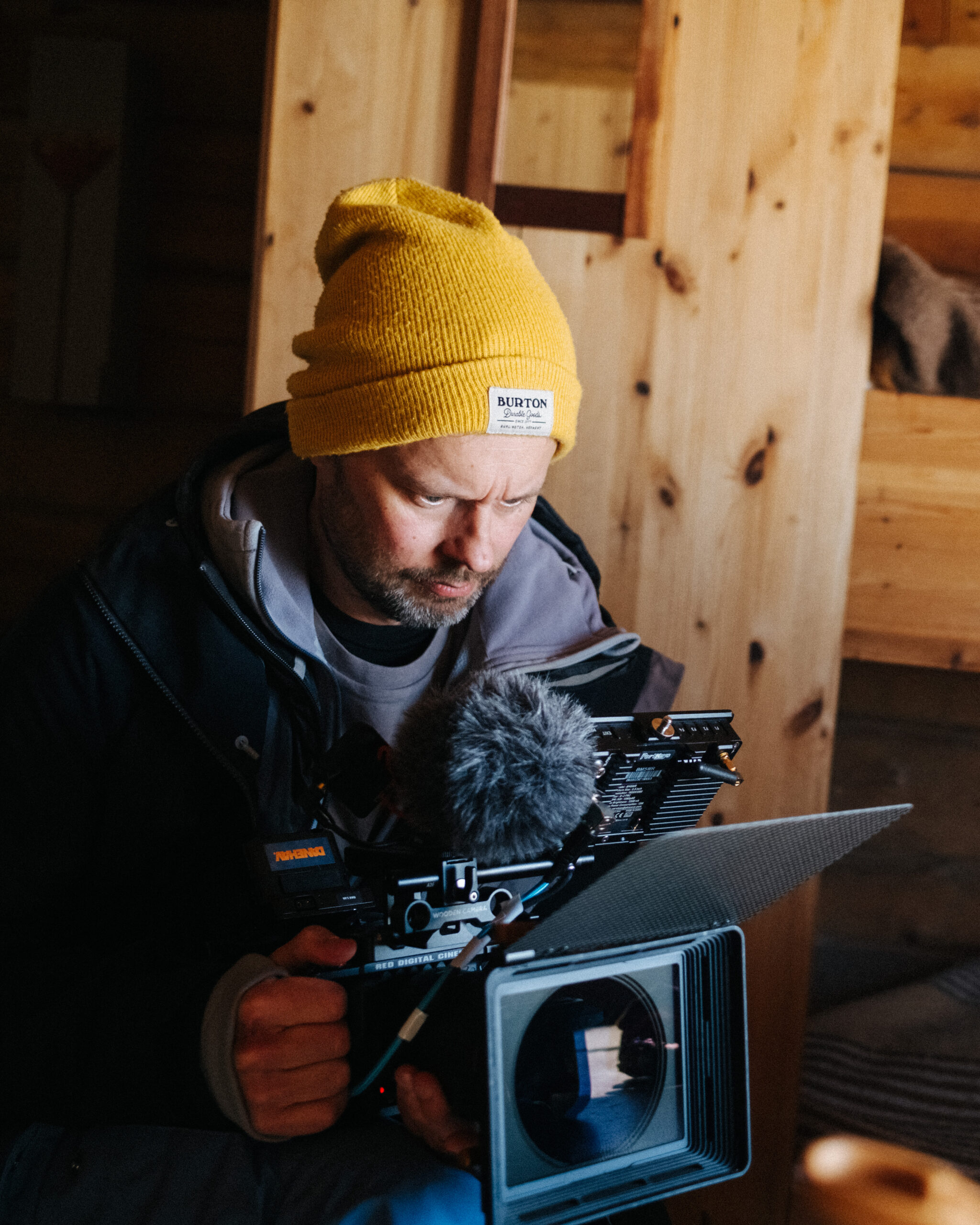 Robin Danehav in a yellow beanie, looking at the screen of a cinema camera in a wooden cabin.